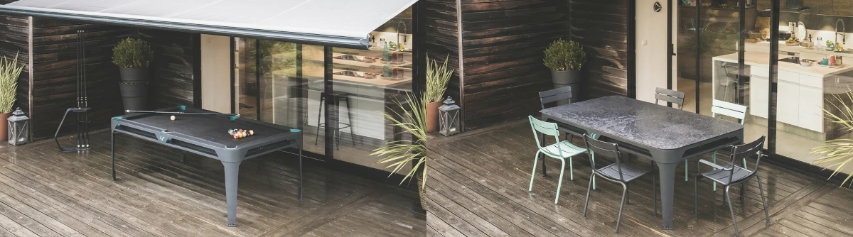 Table de billard convertible en table à manger installée sur une terrasse en bois moderne, avec store extérieur, chaises colorées, plantes décoratives et cuisine visible derrière de grandes baies vitrées.