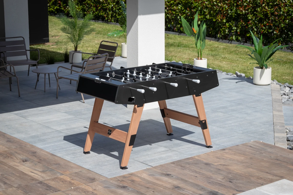 Baby-foot d’extérieur Cornilleau noir avec pieds en bois installé sur une terrasse